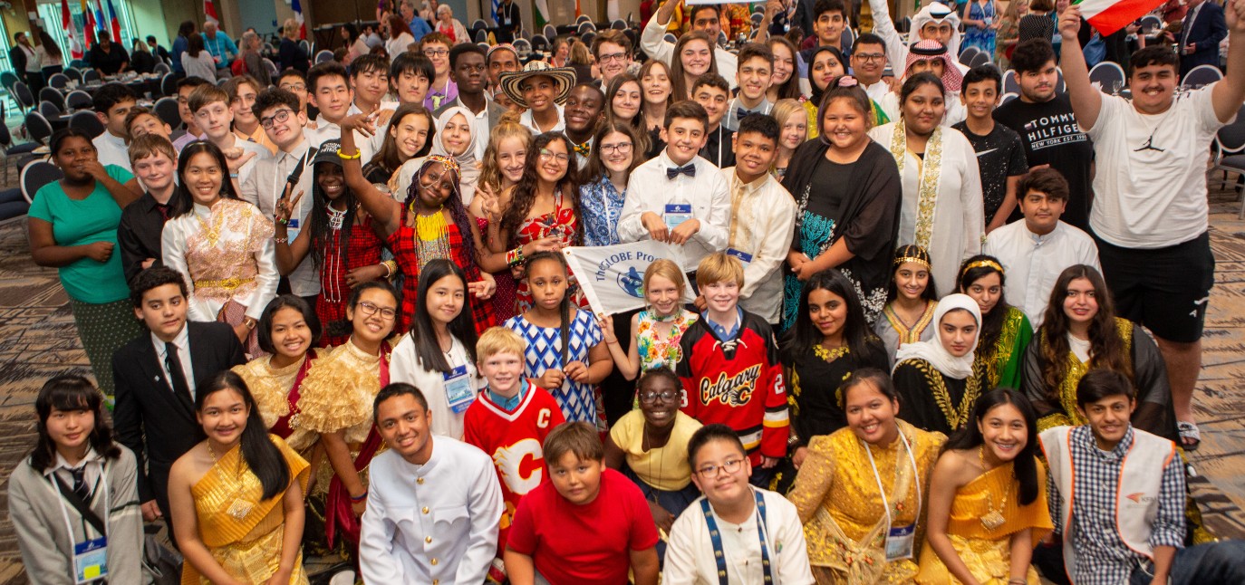 About 50 students are smiling and posing. One student is holding a country flag and another is holding a GLOBE Program flag. 
