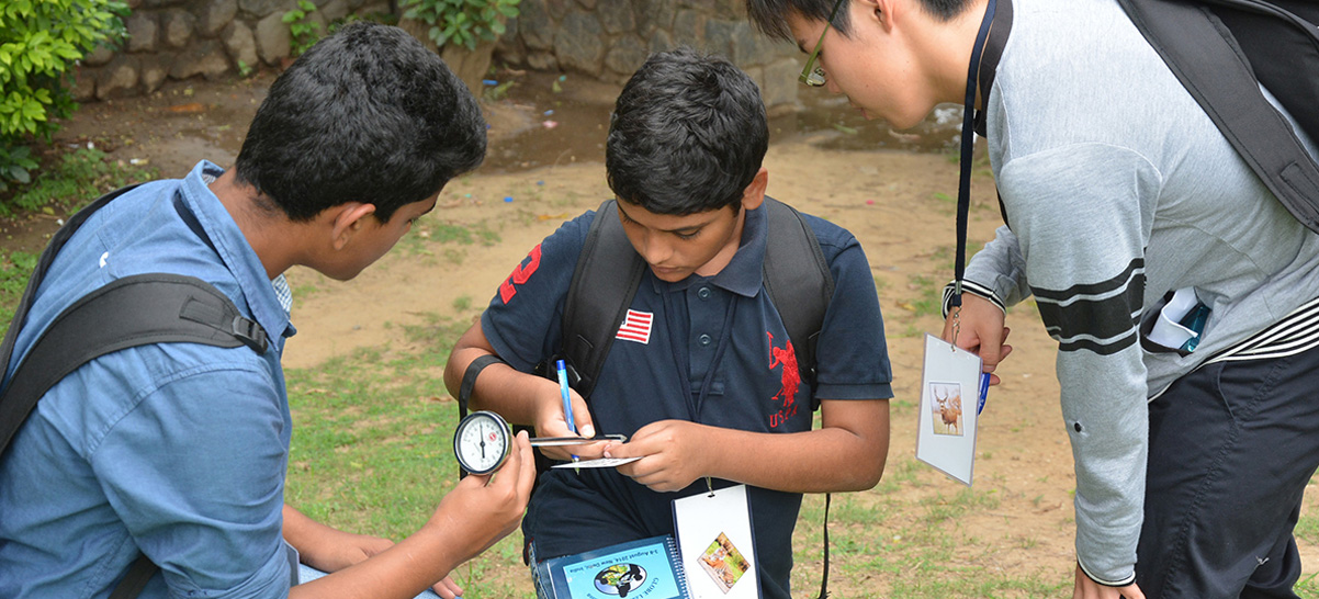 Three boys outside, standing and wearing backpacks. They are all looking at a data sheet and holding scientific instruments. The boy holding the sheet also has a workbook and pen.